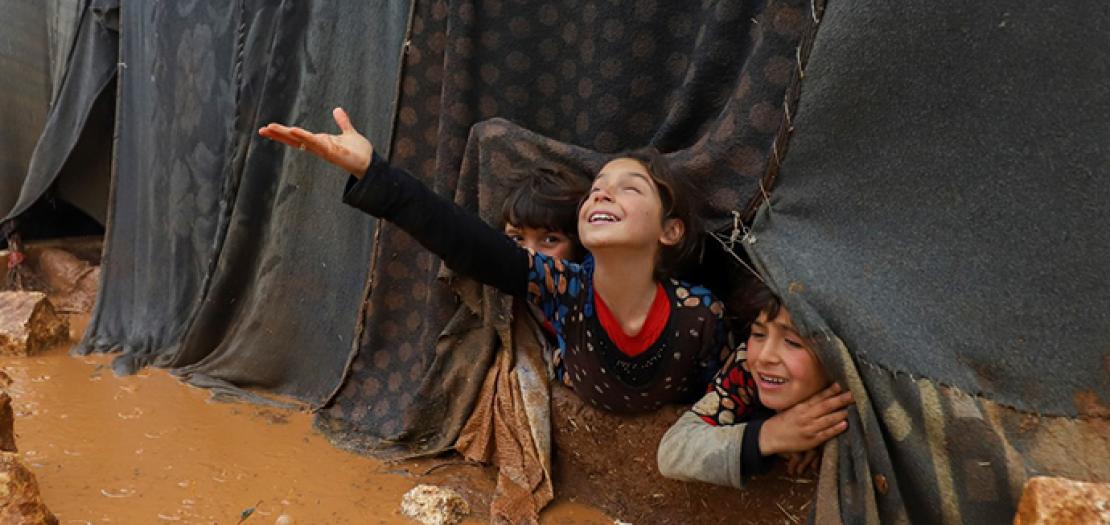 رغم كل الألم، طفلة سورية تبتسم وتمدّ يدها للمطر. الصورة من مخيم أم جرن في ضواحي إدلب بسورية بعد هطول المطر الذي تسبب بسيول وغرق مئات الخيام وتشرّد سكّانها (أ ف ب)